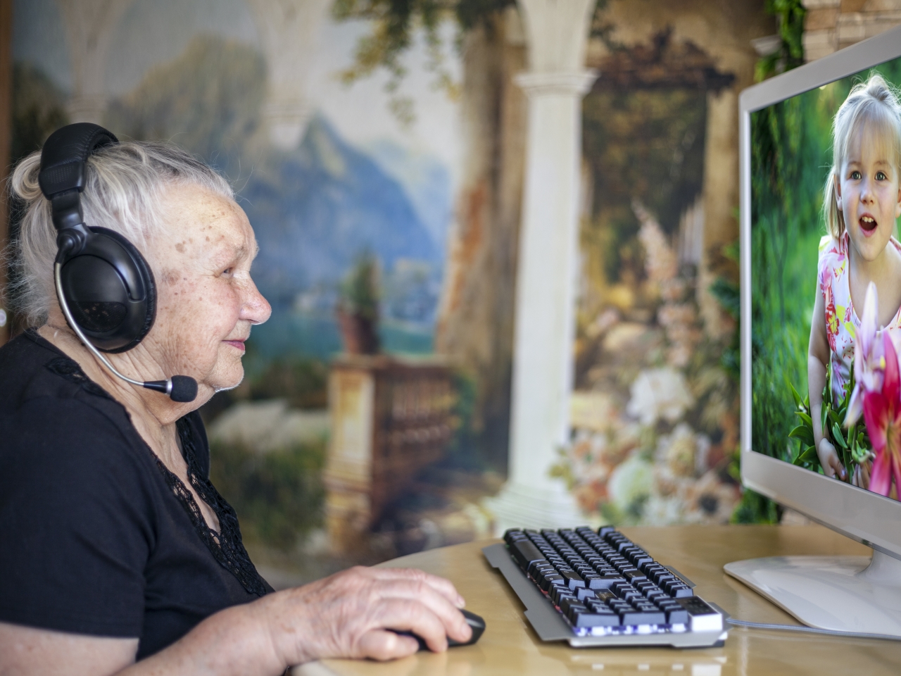 Eine ältere Frau sitzt mit einem Kopfhörer vor einem Bildschirm auf dem ihre Enkeltochter zu sehen ist.