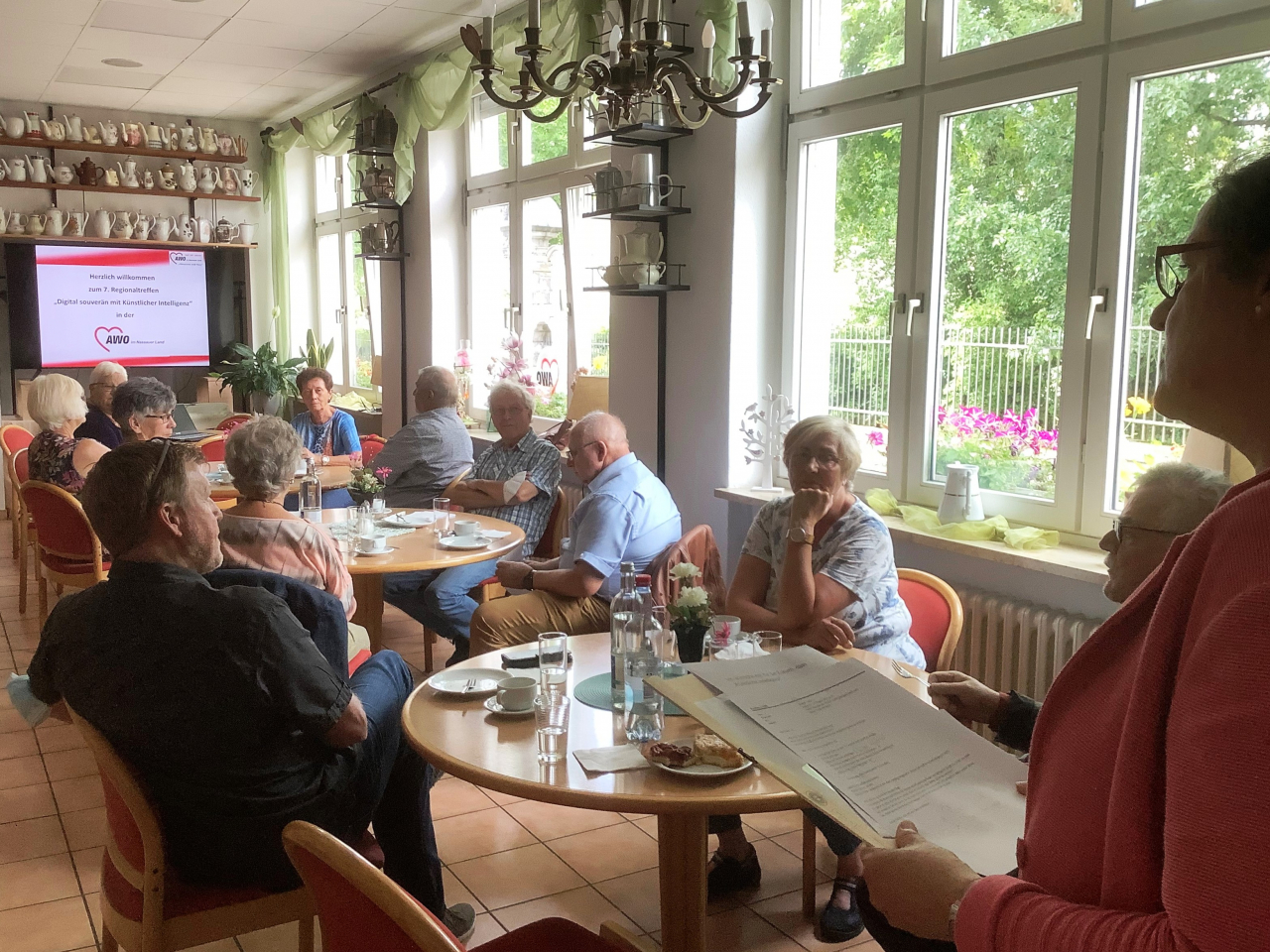 Gruppe älterer Menschen trinkt gemütlich Kaffee, im Hintergrund steht ein Smart-TV