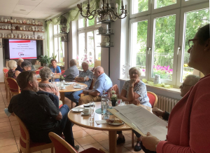 Gruppe älterer Menschen trinkt gemütlich Kaffee, im Hintergrund steht ein Smart-TV